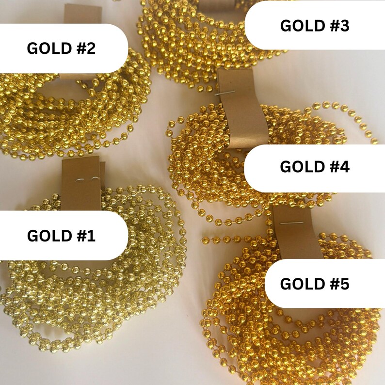 May include: Five coils of metallic gold ball chains, each secured with a tan paper tag. The coils are labeled with black text as GOLD #1, GOLD #2, GOLD #3, GOLD #4, and GOLD #5. The chains consist of small, uniform spherical beads linked together.