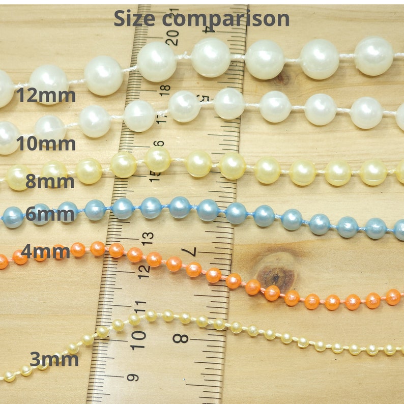 May include: Six strands of round beads in varying sizes and colors, including white, yellow, blue, orange, and cream, are arranged horizontally across a wooden ruler. The text labels 12mm, 10mm, 8mm, 6mm, 4mm, and 3mm indicate the bead diameters.