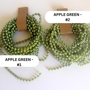 May include: Two coiled strands of apple green ball chain, each secured with a brown paper band. The left coil is labeled APPLE GREEN - #1 and the right coil is labeled APPLE GREEN - #2. The chains feature small, uniform, glossy spherical beads.