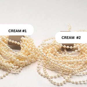 May include: Two bundles of cream-colored pearl bead strands, each wrapped around a brown cardboard holder. The left bundle is labeled CREAM #1 and the right bundle is labeled CREAM #2. The beads are small, round, and have a glossy finish.