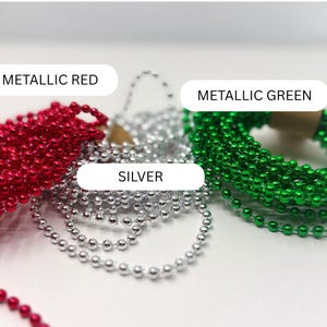 May include: Three coiled strands of metallic ball chain beads in red, silver, and green. Each strand features small, uniform spherical beads connected by thin metal links. The image includes text labels identifying the colors as METALLIC RED, SILVER, and METALLIC GREEN.