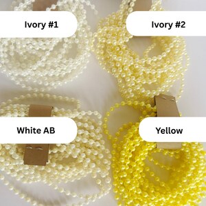 May include: Four coiled strands of faux pearl beads, each secured with a brown paper tag. The colors are labeled as Ivory #1, Ivory #2, White AB, and Yellow. Each strand consists of small, round, uniform beads linked together in a continuous chain.