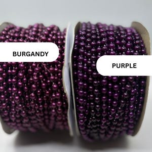 May include: Two spools of ball chain, one labeled BURGANDY and one labeled PURPLE. The burgundy chain features a deep, metallic red-toned finish, while the purple chain displays a vibrant, glossy violet hue. Both are wound on cardboard spools.