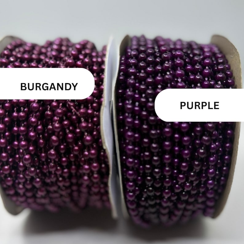 May include: Two spools of ball chain, one labeled BURGANDY and one labeled PURPLE. The burgundy chain features a deep, metallic red-toned finish, while the purple chain displays a vibrant, glossy violet hue. Both are wound on cardboard spools.