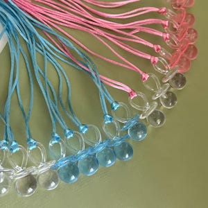 May include: A collection of baby pacifiers with clear, round nipples and blue or pink cords. The pacifiers are made of clear plastic and are arranged on a light green surface. The cords are tied to the pacifier's handle.