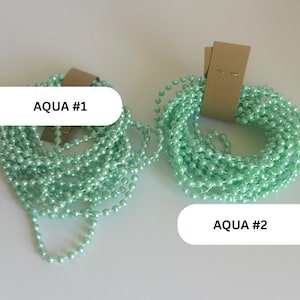 May include: Two coils of aqua-colored ball chain, each secured with a brown cardboard tag. The left coil is labeled AQUA #1 and the right coil is labeled AQUA #2. The chains consist of small, round, uniform beads linked together in a continuous strand.