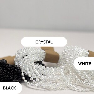 May include: Three strands of ball chain in black, crystal, and white. Each strand is coiled and labeled with the text CRYSTAL, WHITE, and BLACK. The chains consist of small, uniform spherical beads linked together in a continuous line.