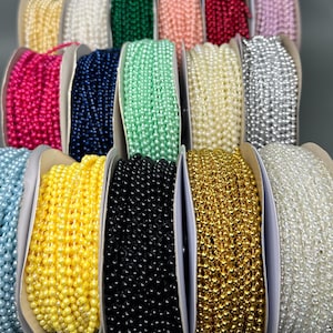 May include: Assortment of spools of pearl-like beads in various colors, including white, black, gold, yellow, pink, blue, green, and red. The beads are arranged in a row, with each spool showing the color of the beads.