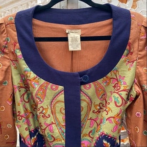 Może przedstawiać: Kurtka w stylu vintage o unikalnym designie. Posiada wz&oacute;r paisley w odcieniach zieleni, r&oacute;żu i pomarańczy, z granatowymi wykończeniami i zapięciem na jeden guzik. Okrągły dekolt i długie rękawy. Wymiary w centymetrach.