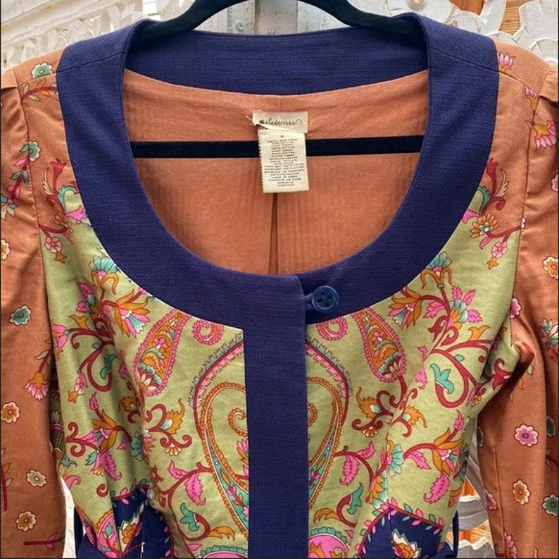 Może przedstawiać: Kurtka w stylu vintage o unikalnym designie. Posiada wz&oacute;r paisley w odcieniach zieleni, r&oacute;żu i pomarańczy, z granatowymi wykończeniami i zapięciem na jeden guzik. Okrągły dekolt i długie rękawy. Wymiary w centymetrach.