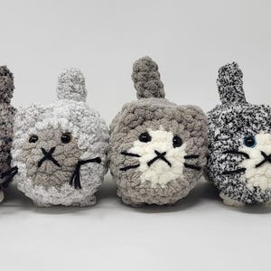 May include: Four gray and white crocheted cat plushies with black eyes and whiskers. The plushies are arranged in a row from left to right.