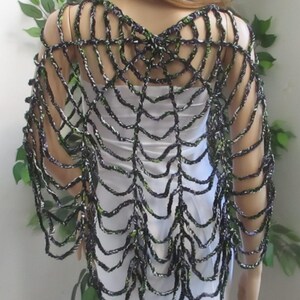 May include: A black and green crocheted shawl featuring an open, web-like pattern that drapes over the shoulders and arms. The garment is constructed from variegated yarn with dark tones and bright green accents, creating a textured, layered look.