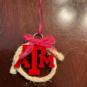 Puede incluir: Adorno de fieltro rojo y blanco con las letras "A" y "M" a cada lado de una gran "T" roja. El adorno está rodeado de una cuerda marrón y tiene un lazo de cinta rojo.