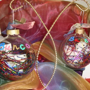 Puede incluir: Dos adornos de vidrio transparente con confeti de colores y las palabras "Bunco" y "B.E.C." escritas en ellos. Los adornos est&aacute;n colgados de un alambre dorado.