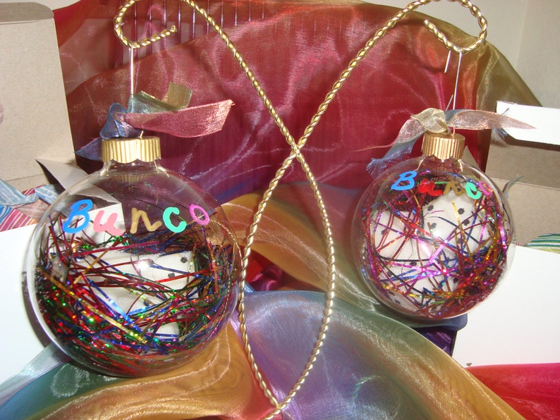 Puede incluir: Dos adornos de vidrio transparente con confeti de colores y las palabras "Bunco" y "B.E.C." escritas en ellos. Los adornos est&aacute;n colgados de un alambre dorado.