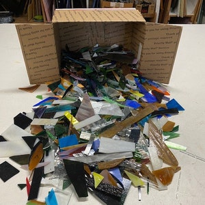 May include: A cardboard box overflowing with a variety of colourful glass pieces, including clear, blue, green, and brown. The glass pieces are of various shapes and sizes, and some are iridescent.
