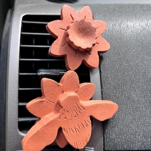 May include: Two terracotta-colored flower-shaped car air freshener clips attached to a vehicle air vent. The top flower has a ruffled center, while the bottom flower features etched petal details. Both items are matte and have a porous texture.