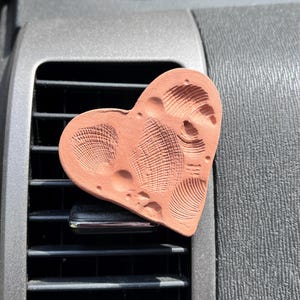 May include: A heart-shaped terracotta-colored air freshener clip featuring embossed seashell patterns on its surface. The object is attached to a black plastic vehicle air vent, displaying detailed textures of various small shells pressed into the clay.