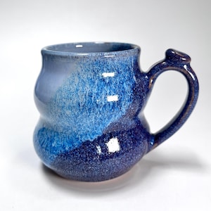 May include: A handmade ceramic mug with a unique, bulbous shape and a dark blue handle. The mug features a gradient of blues, from light to dark, with a speckled texture. The interior is a lighter shade of blue.
