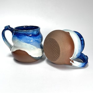 May include: Two handmade ceramic mugs. One upright, the other inverted. Both feature a blue, white, and brown color scheme. The mugs have a unique, organic shape with a handle. The base of the inverted mug has a textured, wavy pattern.