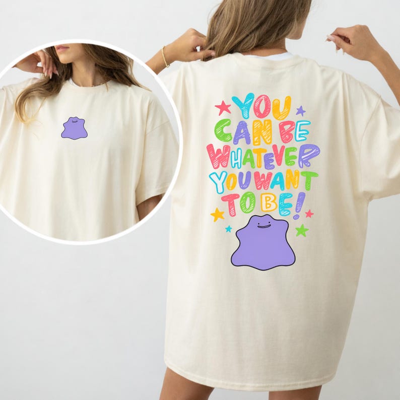 May include: Cream-colored t-shirt featuring a purple cartoon character on the front and back. The back of the shirt has the text "YOU CAN BE WHATEVER YOU WANT TO BE!" in colorful letters, with stars and the cartoon character.