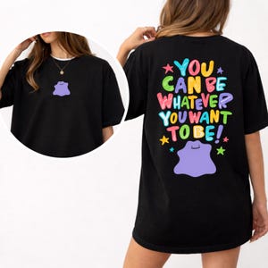 May include: Black t-shirt featuring a purple blob graphic on the front and back. The back of the shirt has the text "YOU CAN BE WHATEVER YOU WANT TO BE!" in colorful letters with stars. The shirt is a comfortable, casual style.