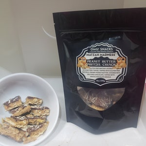 May include: A black resealable bag of OMG! Snacks Matzah Madness Peanut Butter Pretzel Crunch. The bag shows a close-up of the snack with a white chocolate drizzle. The bag is labeled with the product name, ingredients, and weight: 6oz/170g.