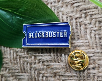 May include: A blue and gold enamel pin shaped like a Blockbuster movie rental ticket. The pin features the word "BLOCKBUSTER" in white lettering. A gold-colored pin back is also visible.