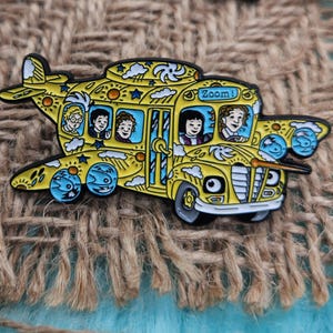 May include: A yellow school bus with wings, painted with clouds, stars, and suns. The bus has a sign that says "Zoom!" and five children are sitting inside. The bus is a pin.