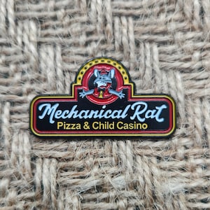 May include: A rectangular pin with the text "Mechanical Rat Pizza & Child Casino." The pin features a cartoon rat character in a red circle with yellow accents. The pin has a black background with red and yellow borders.
