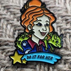 May include: A colorful enamel pin featuring a cartoon woman with red hair, wearing a blue shirt with a star and a crescent moon, and holding a green lizard. The pin has the text "Do it for her" on a blue banner.