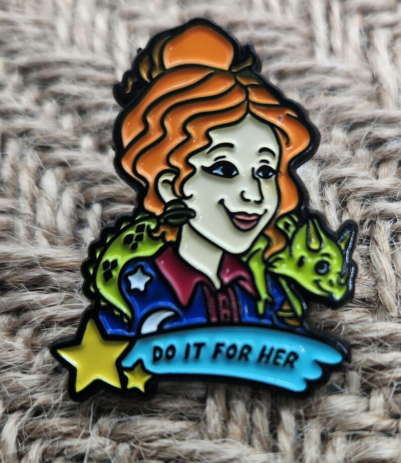 May include: A colorful enamel pin featuring a cartoon woman with red hair, wearing a blue shirt with a star and a crescent moon, and holding a green lizard. The pin has the text "Do it for her" on a blue banner.