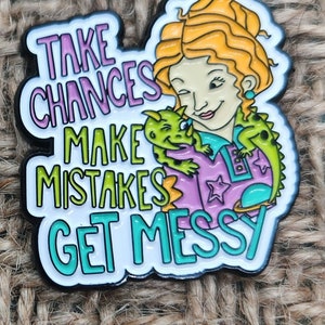 May include: A colorful enamel pin featuring a cartoon woman with red hair holding a green dinosaur. The pin says "Take Chances Make Mistakes Get Messy".