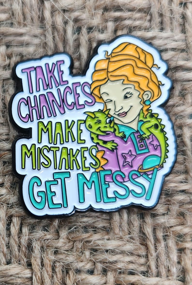 May include: A colorful enamel pin featuring a cartoon woman with red hair holding a green dinosaur. The pin says "Take Chances Make Mistakes Get Messy".