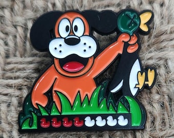 May include: A colorful enamel pin featuring a cartoon dog holding a black duck. The dog is orange and white with black ears. The pin includes green grass and small red and white ducks at the bottom.