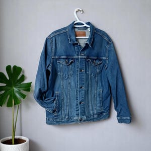 May include: A blue denim jacket with a classic design, featuring a button-up front, chest pockets, and side pockets. The jacket is hanging on a white hanger against a gray wall. The jacket has a brown label on the back.