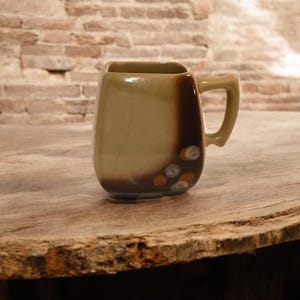 May include: A ceramic mug with a square top and a rounded body. The mug is primarily tan with a dark brown accent on one side and small, decorative white and orange circles near the base. It has a curved handle.