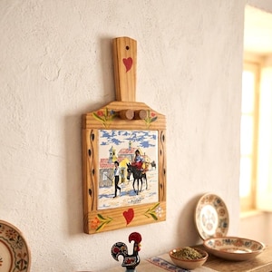 May include: A wooden wall-mounted rack featuring a central ceramic tile with a painted scene of a person leading a donkey through a village. The light-coloured wood frame is decorated with red hearts and floral motifs, and includes two small wooden pegs.