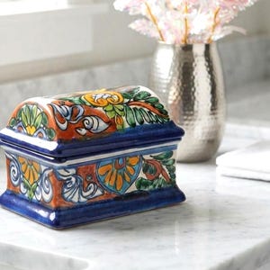 May include: A rectangular ceramic box with a curved lid, featuring a hand-painted floral and scroll pattern in vibrant shades of orange, yellow, green, and blue. The base and edges of the lid are finished in a solid, glossy deep blue glaze.