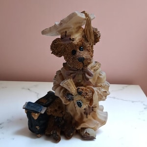 May include: A brown teddy bear figurine wearing a graduation cap and gown, holding a diploma. The bear is seated next to a black and blue book. The figurine has a detailed design and is on a white surface.
