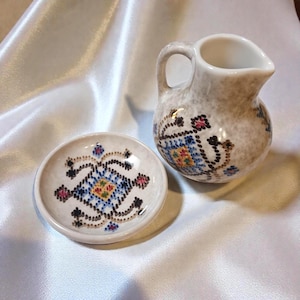 May include: A small, off-white ceramic creamer pitcher and matching saucer. Both pieces are decorated with a repeating pattern of blue, yellow, pink, and black geometric shapes. The pitcher has a handle and a spout.