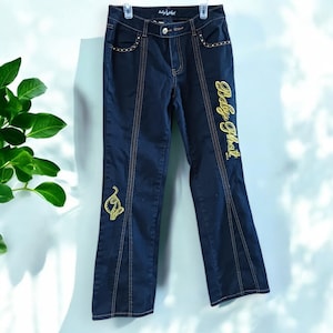 Może przedstawiać: Jeansy z ciemnego denimu z haftem w kolorze złotym, na którym widnieje napis "Baby Phat".