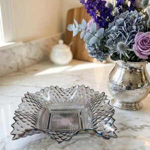 Puede incluir: Una fuente cuadrada de vidrio transparente con un diseño de diamantes y bordes festoneados. La fuente está sobre una encimera de mármol junto a un jarrón plateado lleno de flores moradas y azules. Una tabla de cortar de madera y un azucarero blanco están en el fondo.
