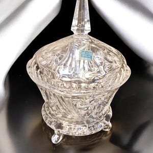 May include: A clear glass candy dish with a lid and a floral design. The dish has three feet and is sitting on a white satin fabric.