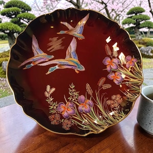 May include: A decorative burgundy plate with a scalloped edge and gold trim. The plate features three flying ducks, purple irises, and gold accents. The plate has a Japanese aesthetic.