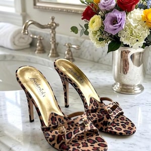 May include: A pair of high-heeled sandals with a leopard print pattern. The sandals have a gold-colored insole and the brand name "Fredericks of Hollywood" is visible. The heels are approximately 4 inches tall.