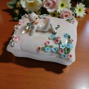 Puede incluir: Caja de baratijas de cerámica blanca con una figura reclinada. La figura tiene una gorra y detalles verdes, con adornos florales rosas y azules. La caja está sobre una superficie de madera, con flores en el fondo.