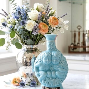 Op de afbeelding: Een lichtblauwe, antieke Gemini-fles met reliëf van cherubijnen en de tekst "Rare Antique". Een boeket witte rozen, oranje ranonkels en blauwe distels staat in een zilveren vaas. De scène speelt zich af op een wit aanrecht.