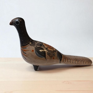 Könnte beinhalten: Keramikvogel-Figur mit langem Schwanz und schlankem Hals. Der Vogel hat einen dunkelbraunen Kopf und Hals, mit einem beigefarbenen Körper, der mit Blumen- und Blattmustern in Braun-, Orange- und Blautönen verziert ist. Der Vogel steht auf einer Holzoberfläche.