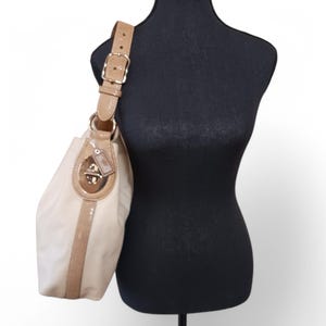 Pode incluir: Uma bolsa Coach de couro bege com fecho de trava dourado e uma alça de couro marrom. A bolsa tem uma faixa branca na frente.